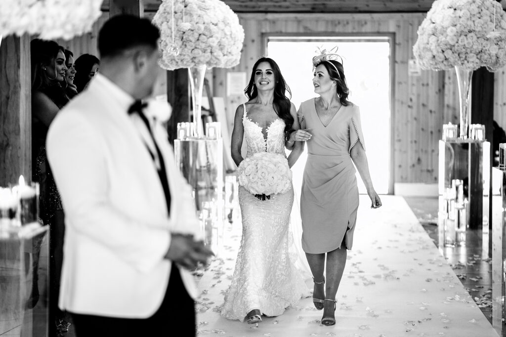 Black and white photo of Gemma walking down the aisle arm in arm with her mum, Tyrone watching in the foreground, Merrydale Manor ceremony room