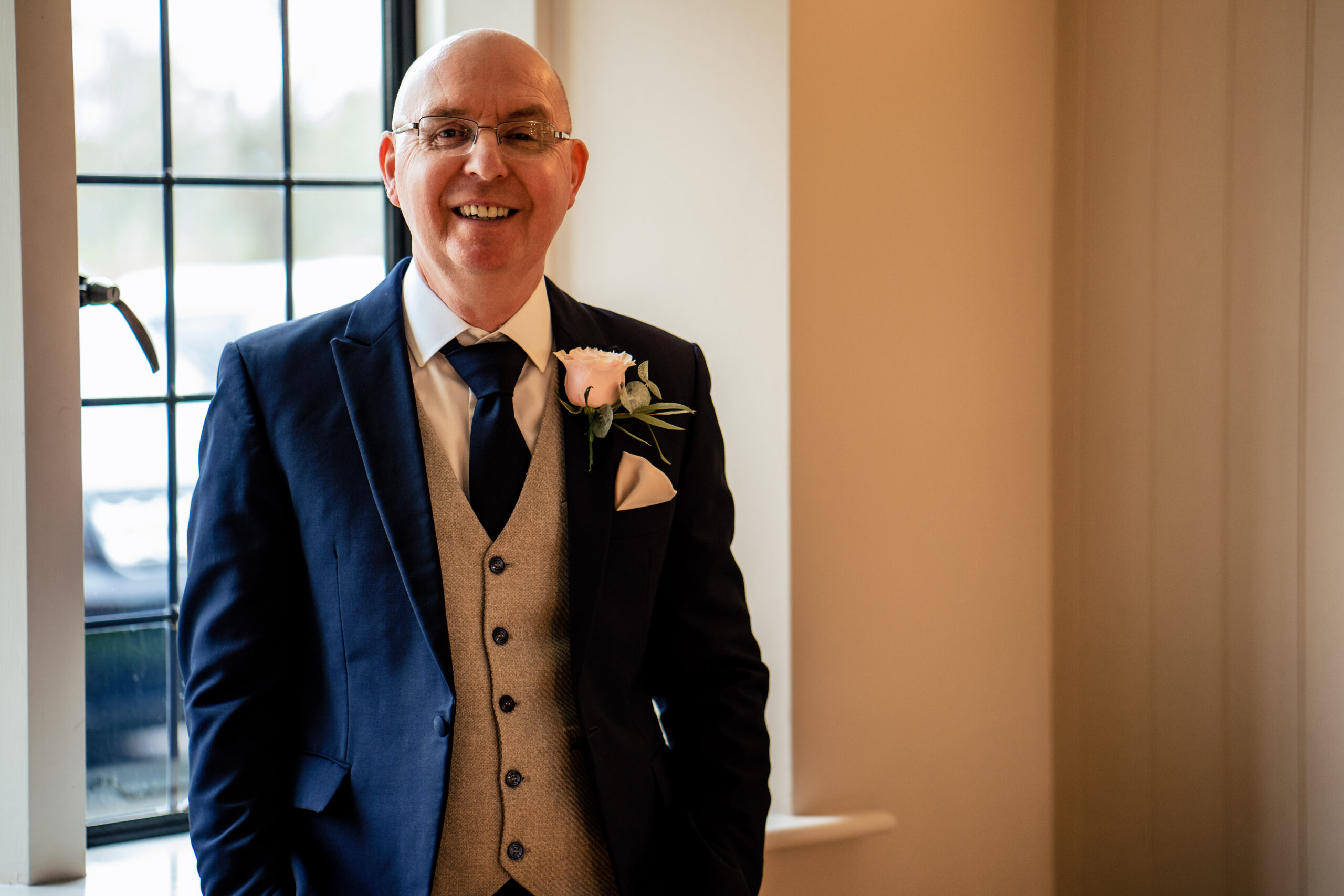 Bethany's dad smiling by a window in his navy suit and pink rose buttonhole, waiting to see her