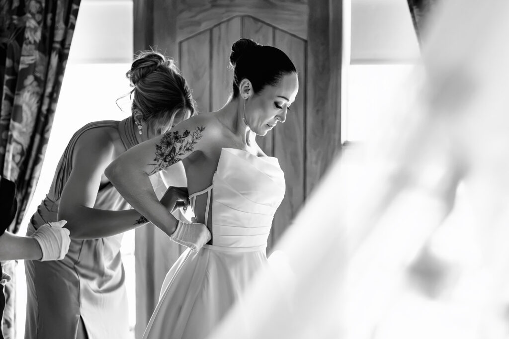 Black and white of hands fastening the back of Kim's strapless dress by the window, Nottingham wedding photographer