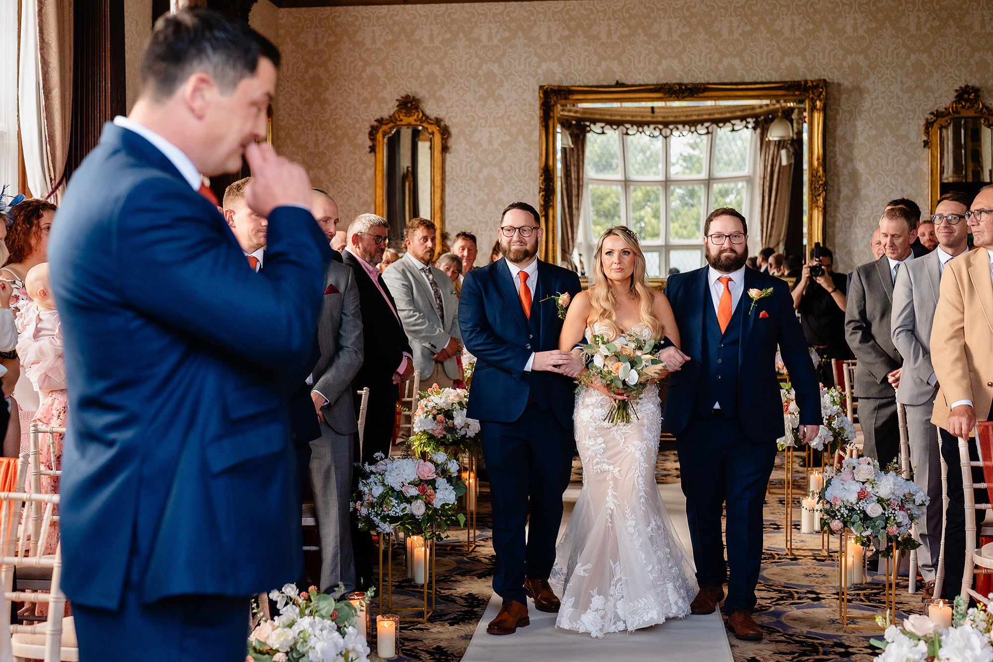 The groom wiping his eye in the foreground as the bride walks down the aisle behind him at Crewe Hall