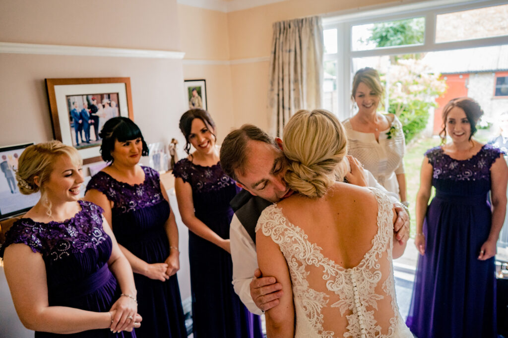 Bride hugged by her dad as bridesmaids in purple dresses look on, Cheshire wedding photographer