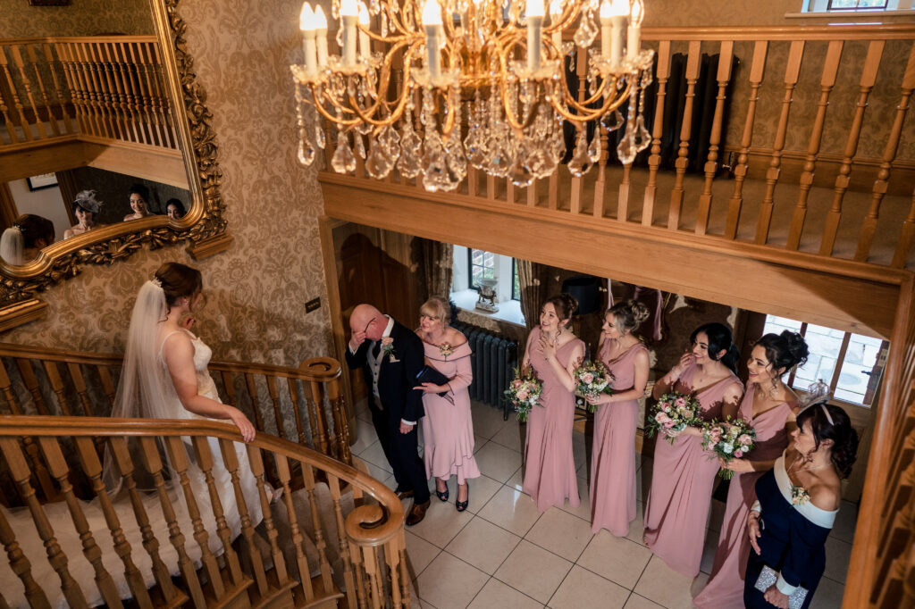 Bethany walking down the staircase at Merrydale Manor towards her dad, mum and bridesmaids waiting below