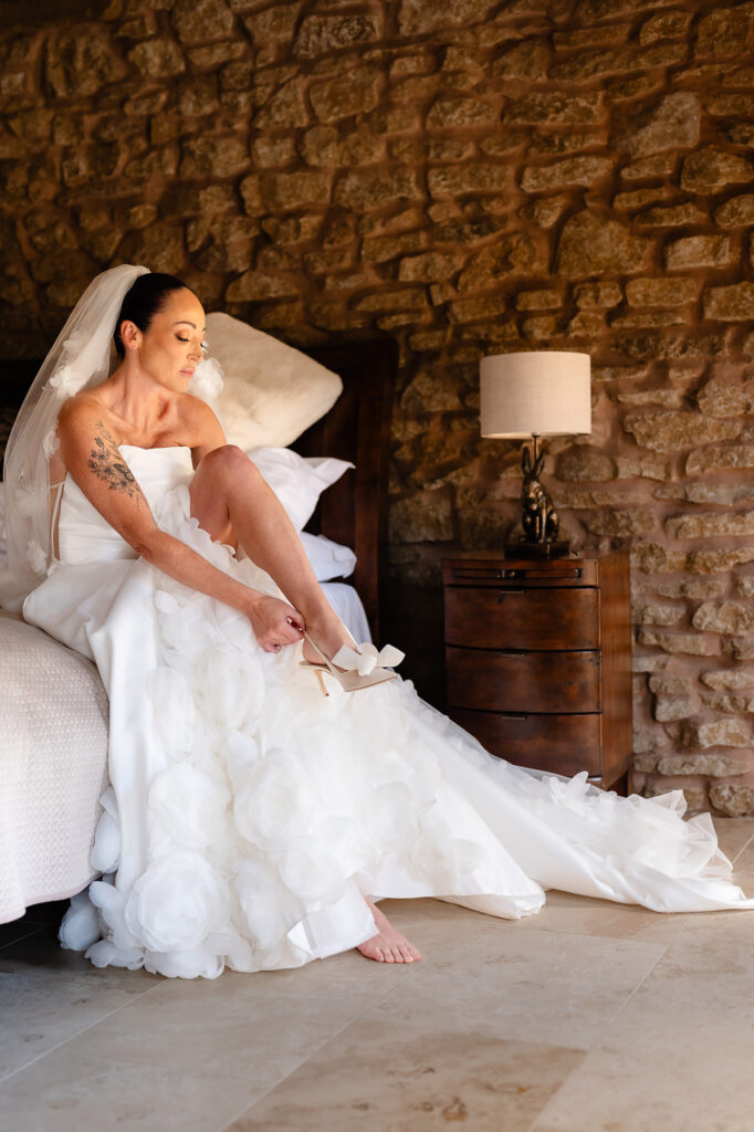 Kim sitting on the edge of the bed in the stone walled stables cottage putting on her shoes, skirt covered in fabric roses, Swancar Farm wedding photographer