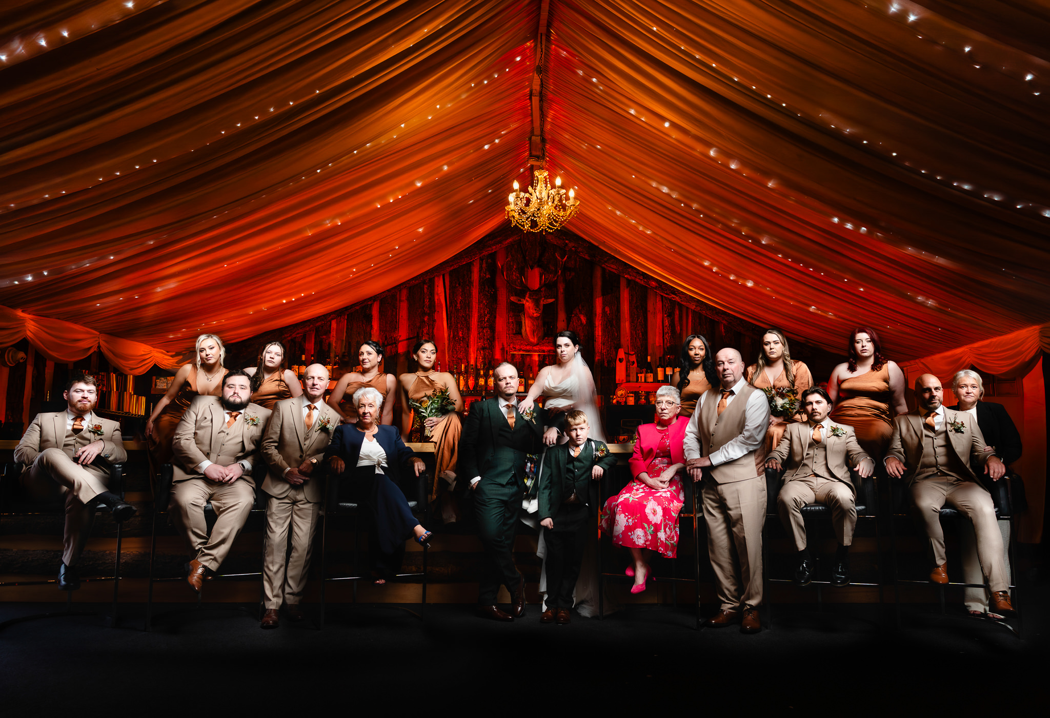 Theatrical group portrait of the whole wedding party posed along the bar under red draped cloth and a chandelier, Heaton House Farm wedding photographer
