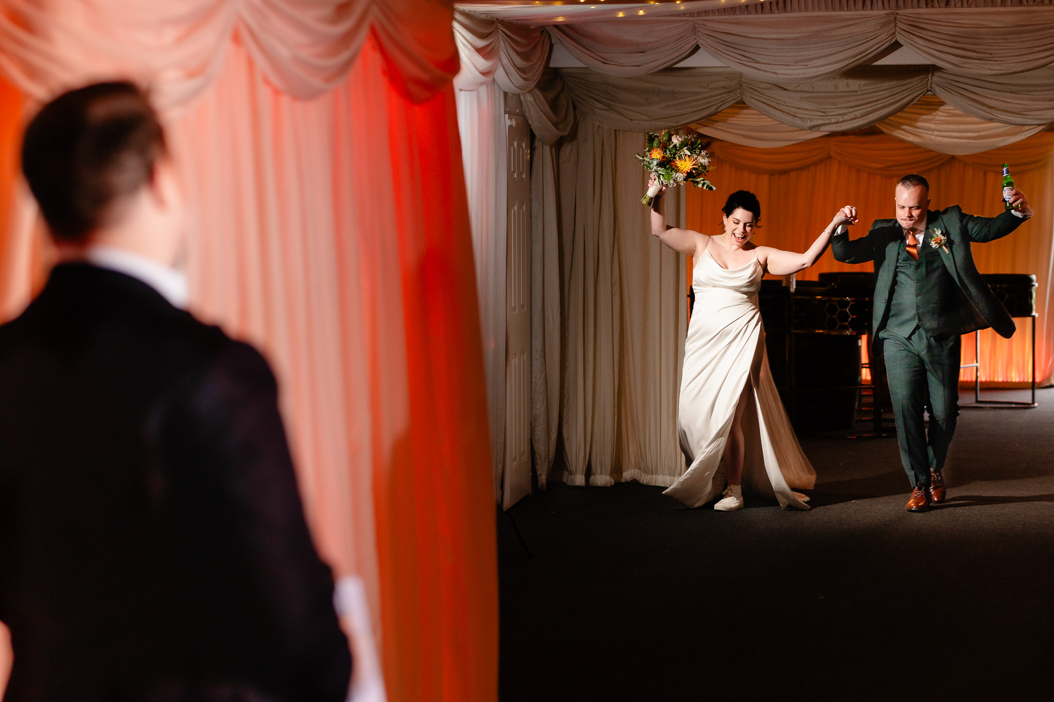 Bride and groom bursting into the draped barn for their wedding breakfast with arms in the air, Heaton House Farm wedding