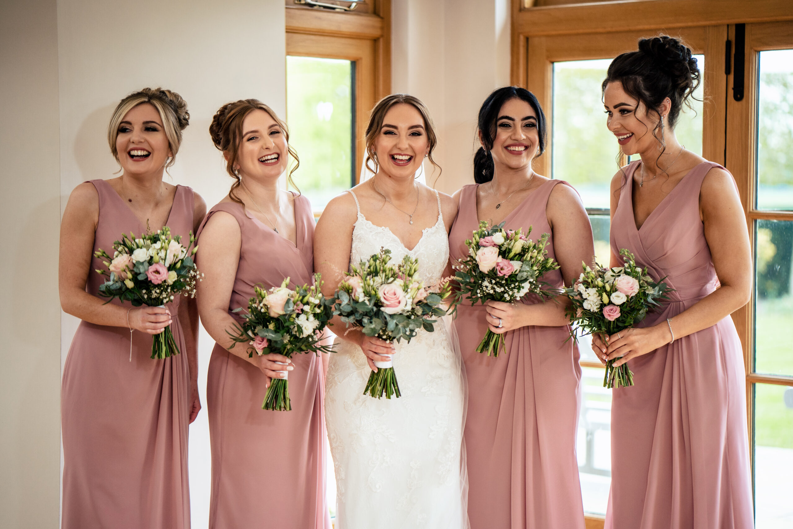 Bethany laughing with her bridesmaids in dusky pink dresses, all holding pink rose bouquets