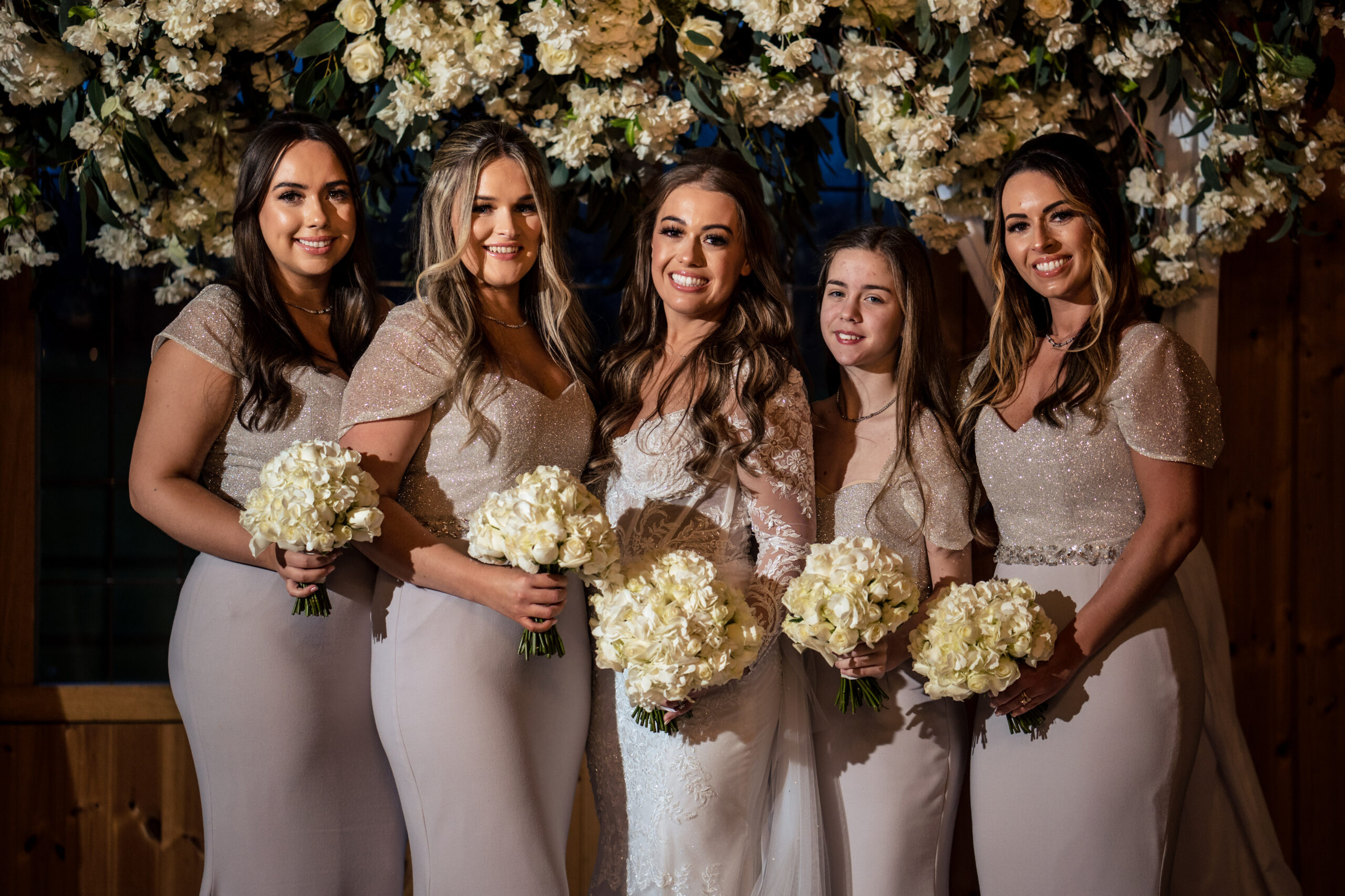 Abbie laughing with her bridesmaids holding white bouquets under the flowers at Merrydale Manor