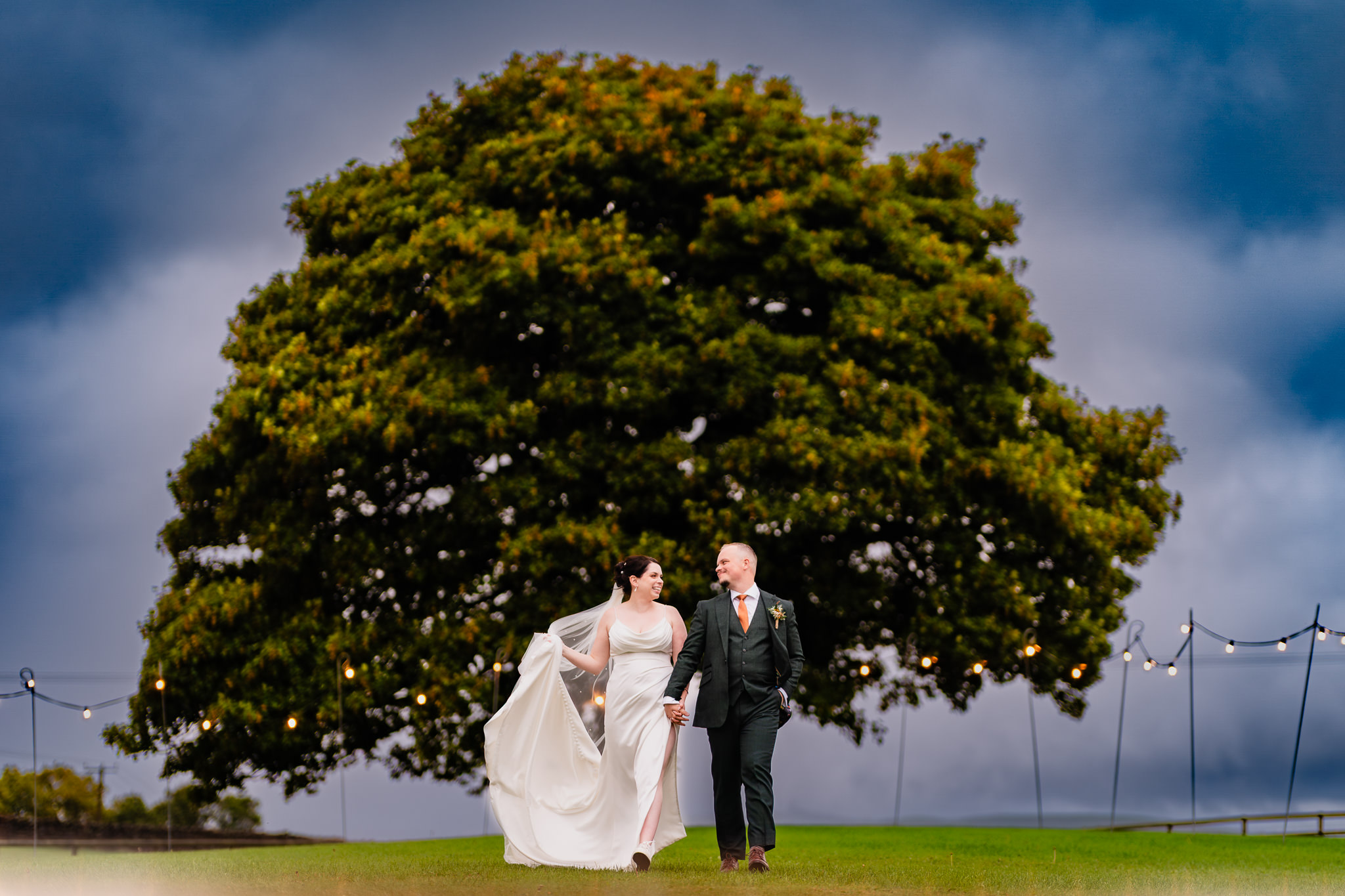 Bride and groom walking together under the festoon lit oak tree in the field at Heaton House Farm, Cheshire wedding photographer