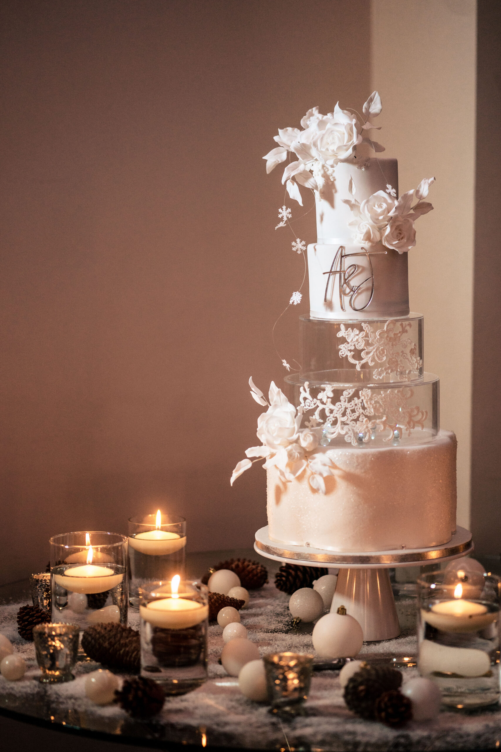 Tall white Confetti and Crumbs wedding cake with sugar flowers and an A and J monogram among candles and pine cones, Merrydale Manor