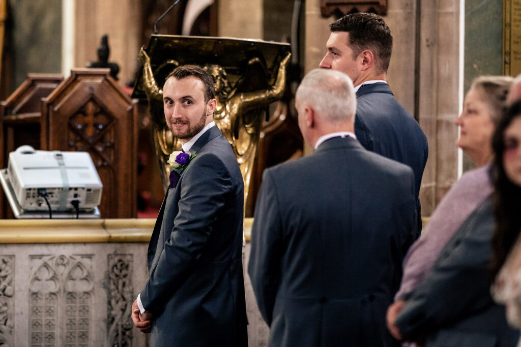 Groom glancing back towards the church door as he waits at the altar, Merrydale Manor wedding photographer