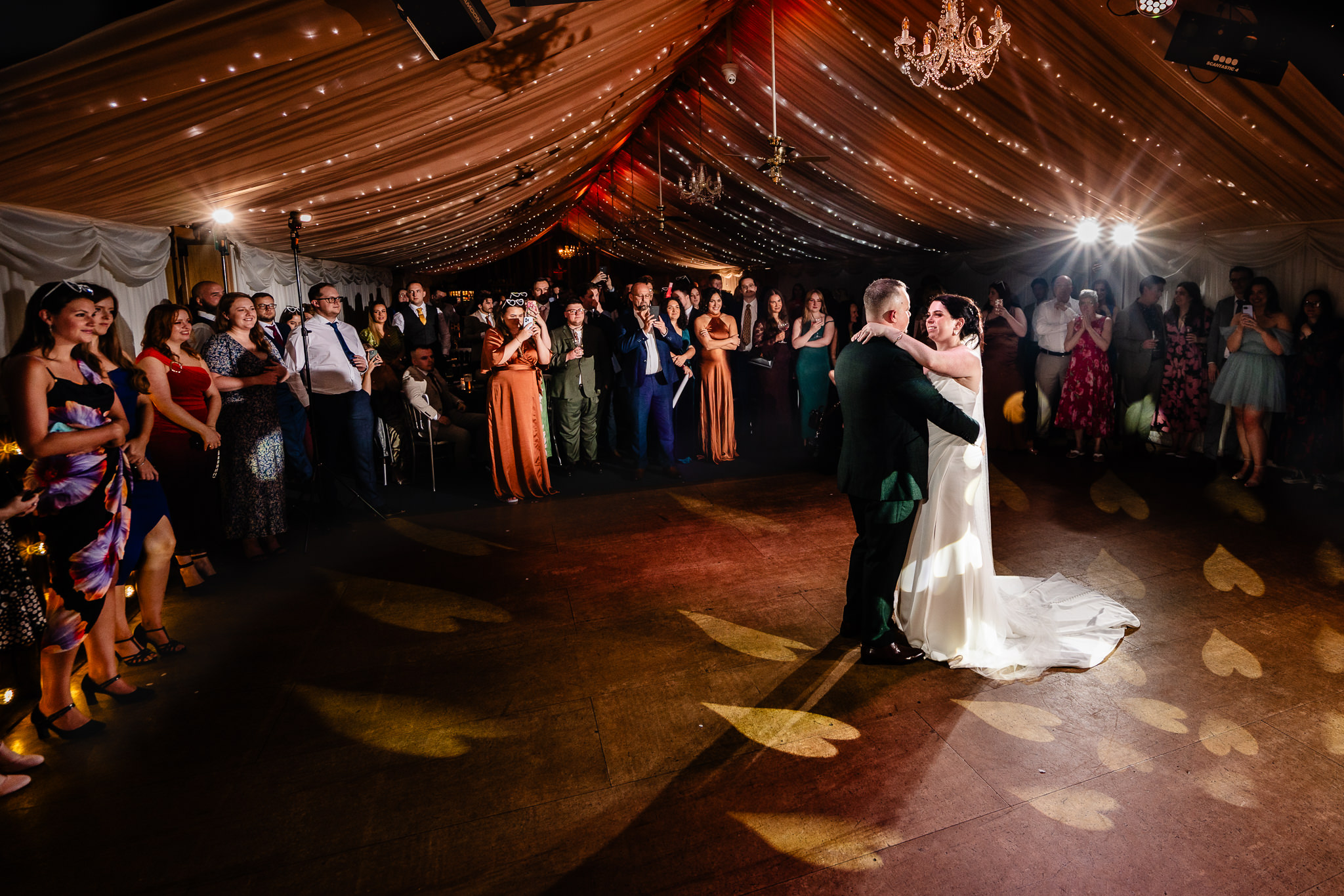 Bride and groom during their first dance in the draped barn surrounded by guests under the chandelier, Heaton House Farm wedding