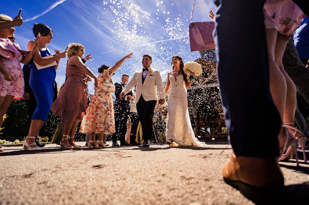 Low angle shot of Gemma and Tyrone walking through a confetti tunnel under a blue sky, Merrydale Manor wedding photographer