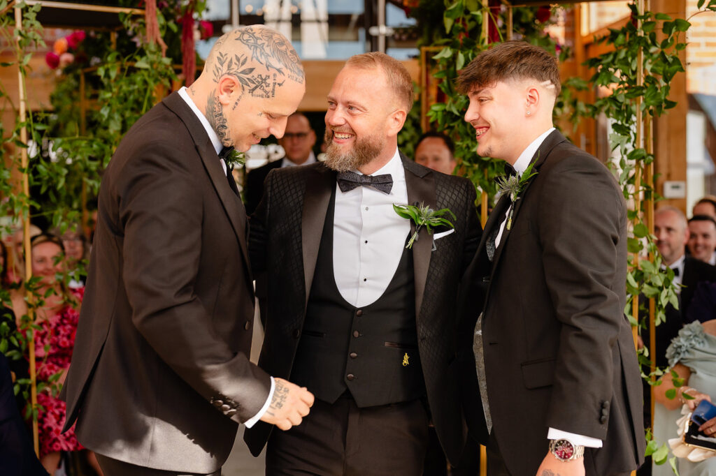 Glynn laughing with two groomsmen at the front of the glass barn before the ceremony, Swancar Farm wedding photographer