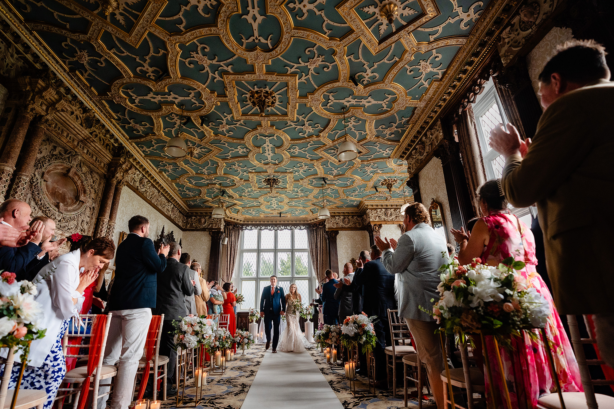 Wide view of the couple walking back down the aisle under the painted and gilded ceiling of the Drawing Room at Crewe Hall
