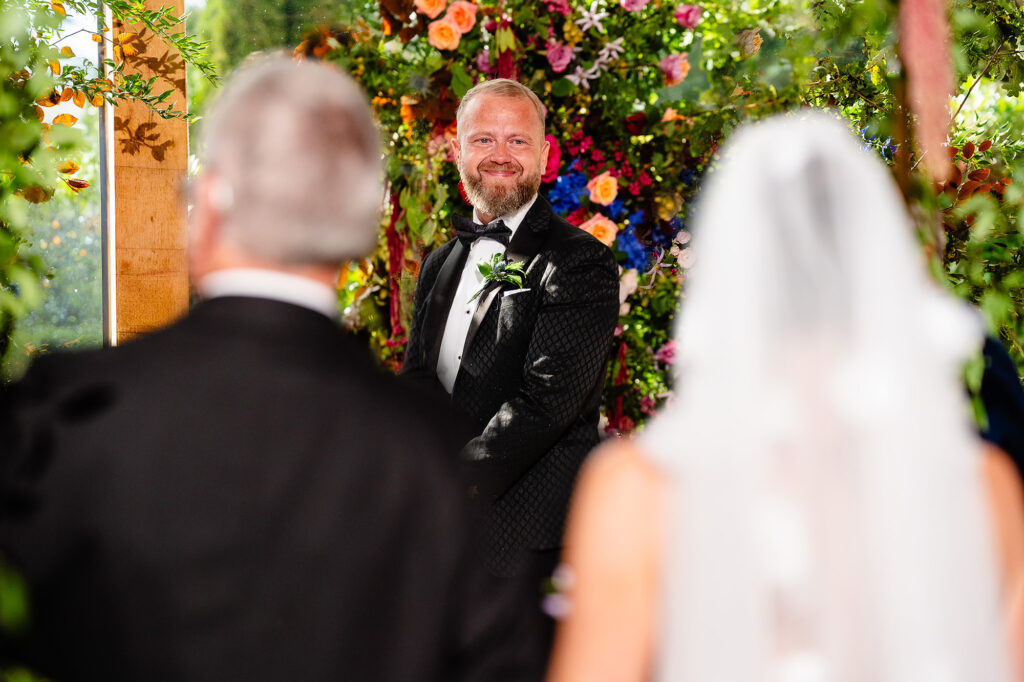 Glynn grinning under the florals as Kim and her dad walk towards him, Swancar Farm wedding photographer