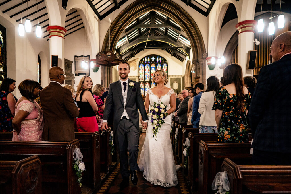 Bride and groom walking back down the aisle as guests stand and cheer, Cheshire wedding photographer