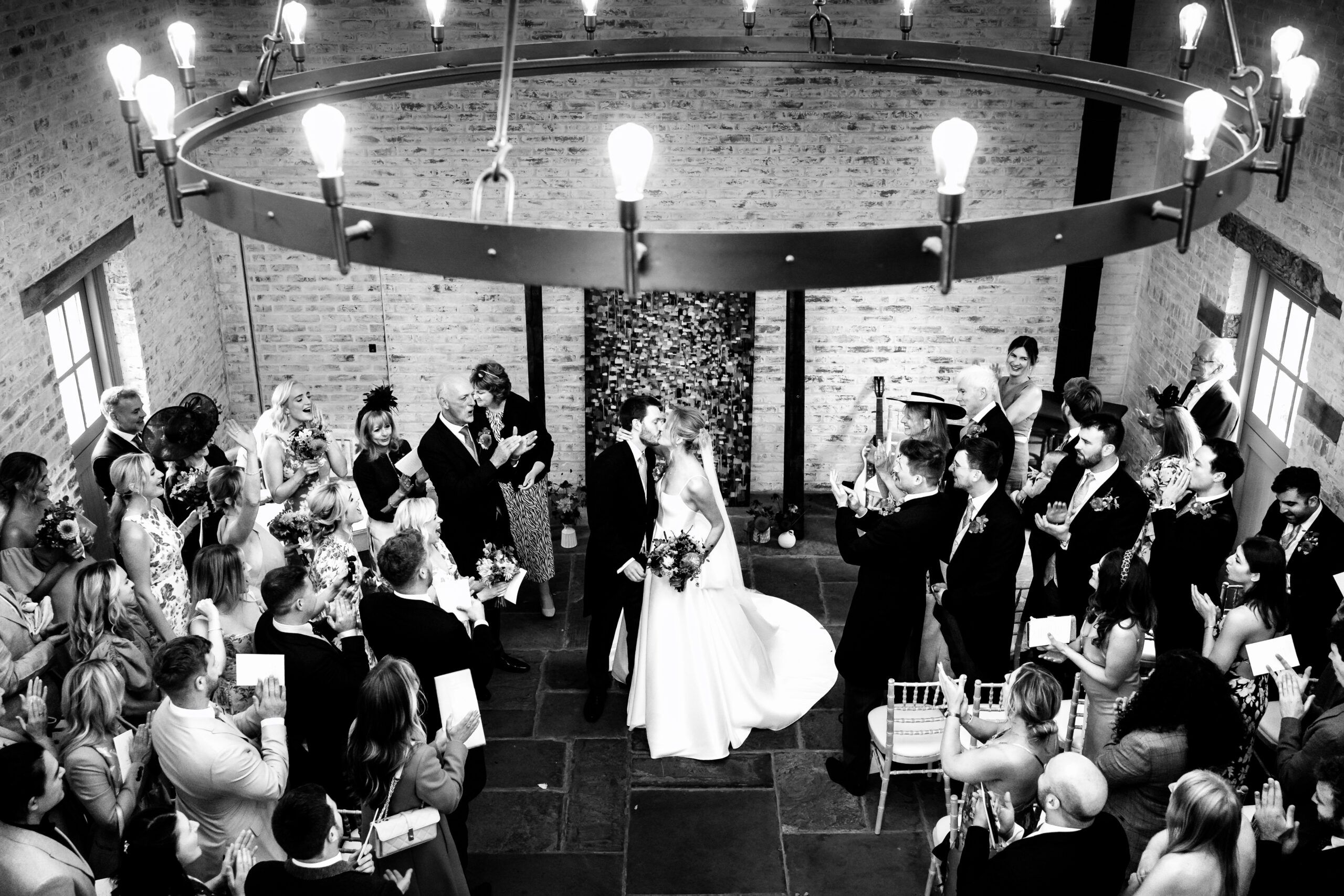 Black and white wide shot of the first kiss in the Coach House at Dorfold Hall as guests stand and applaud