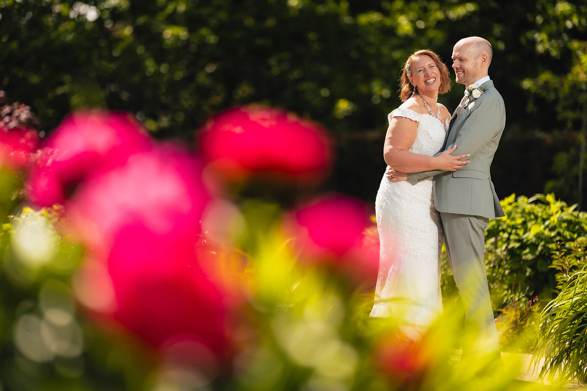 Bride and groom photographed through a foreground of deep red roses in the gardens at Salt House