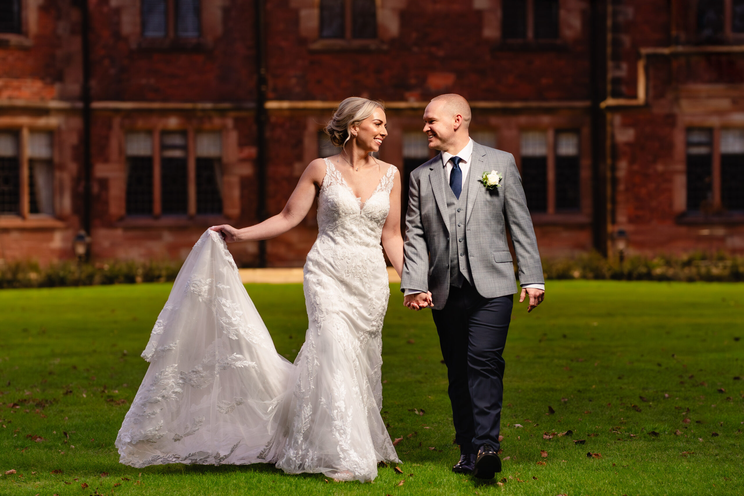 Bride and groom walking across the lawn in front of the red brick hall, her train sweeping behind her, Colshaw Hall Cheshire wedding photographer