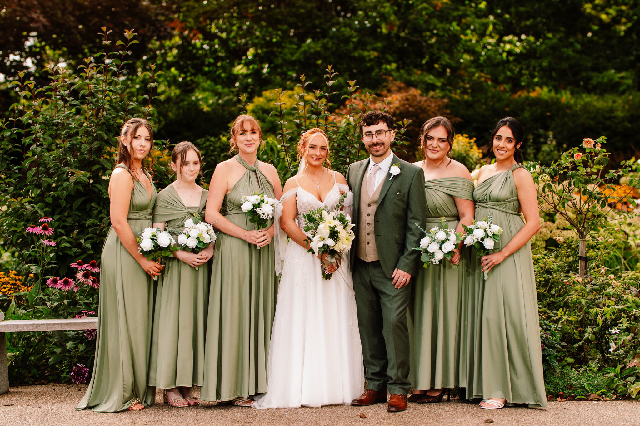 The groom surrounded by the bridesmaids in sage green, all laughing together in the gardens