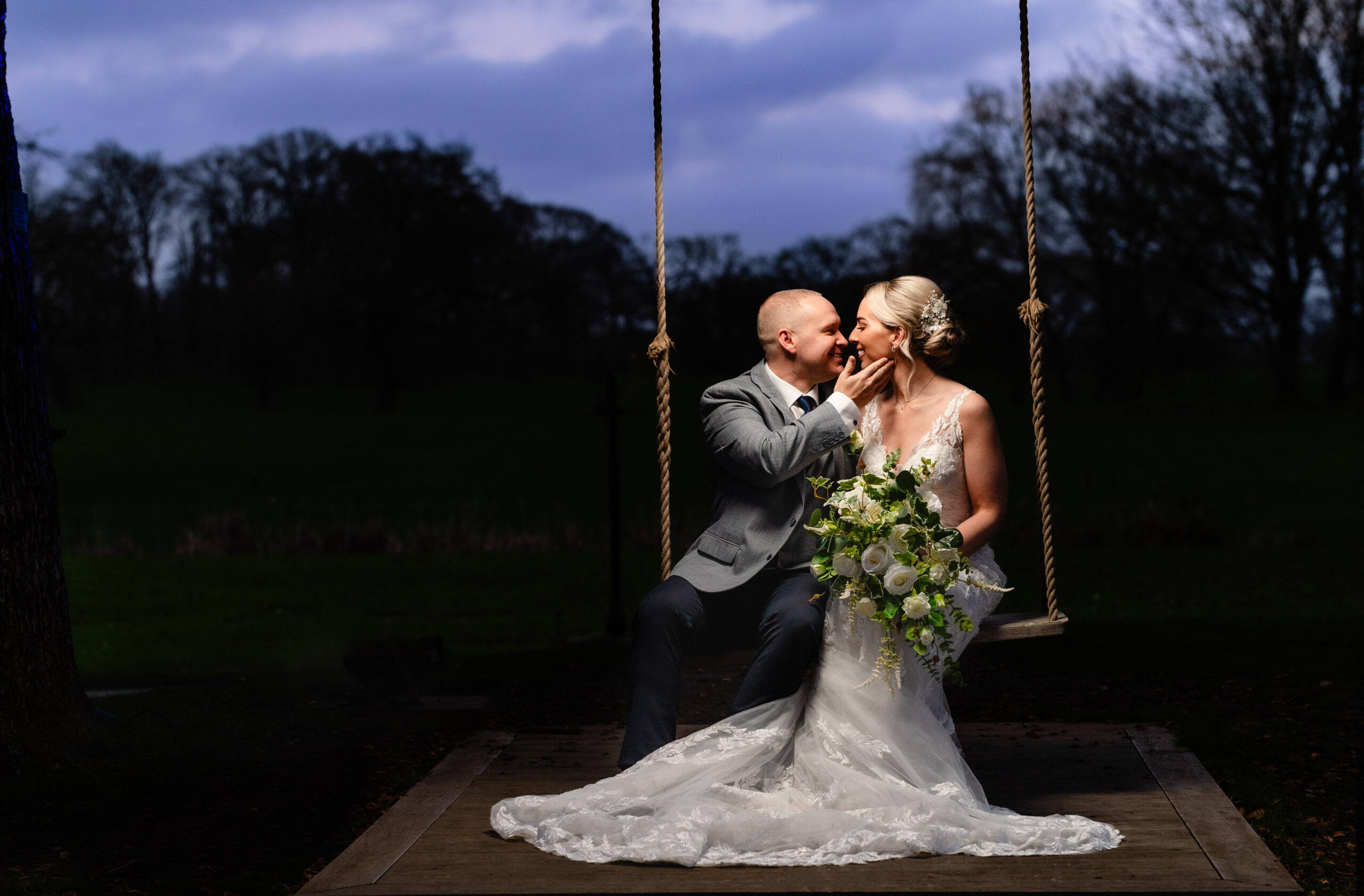 Bride and groom sitting together on the garden swing at dusk with a deep blue winter sky behind them, Colshaw Hall wedding photographer