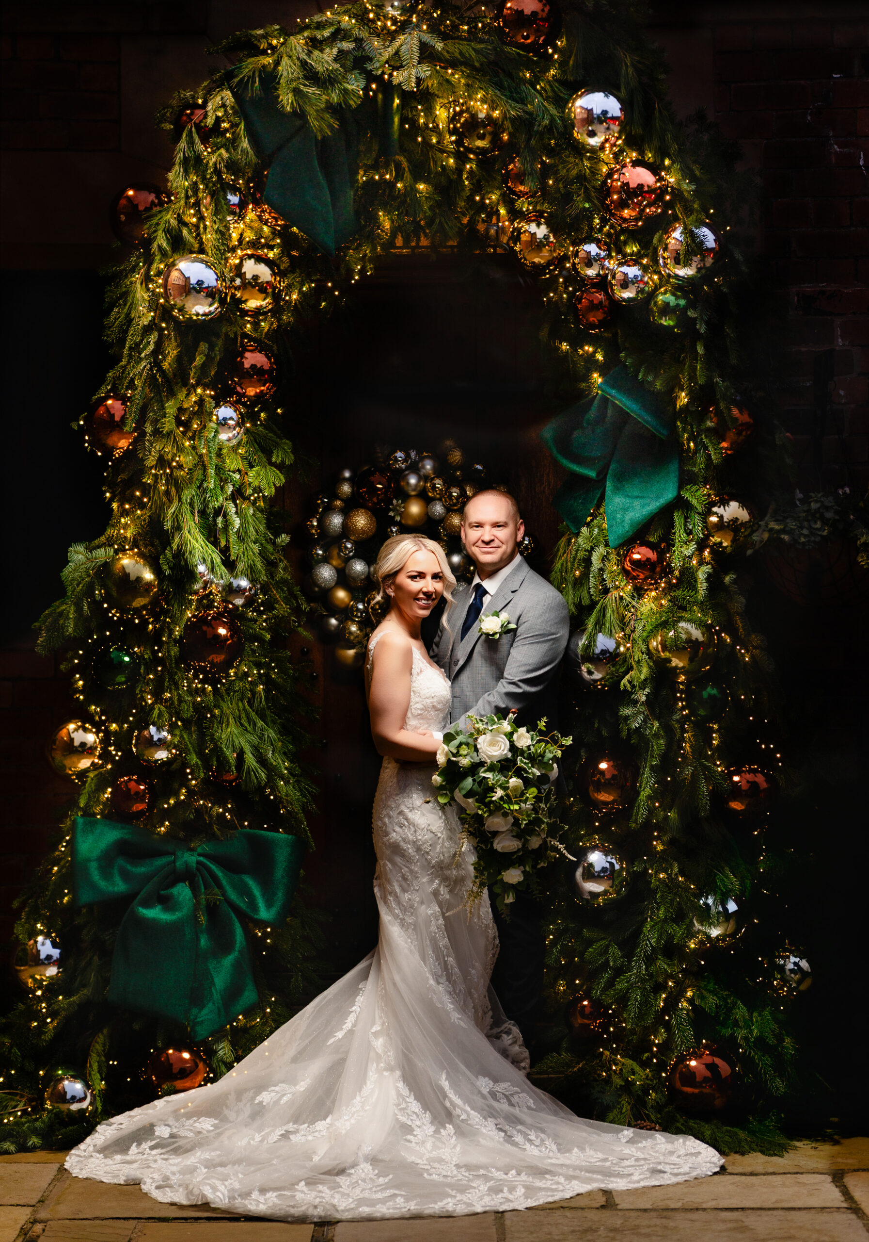 Bride and groom standing inside the giant Christmas wreath arch of foliage, baubles and green bows at Colshaw Hall, Cheshire winter wedding