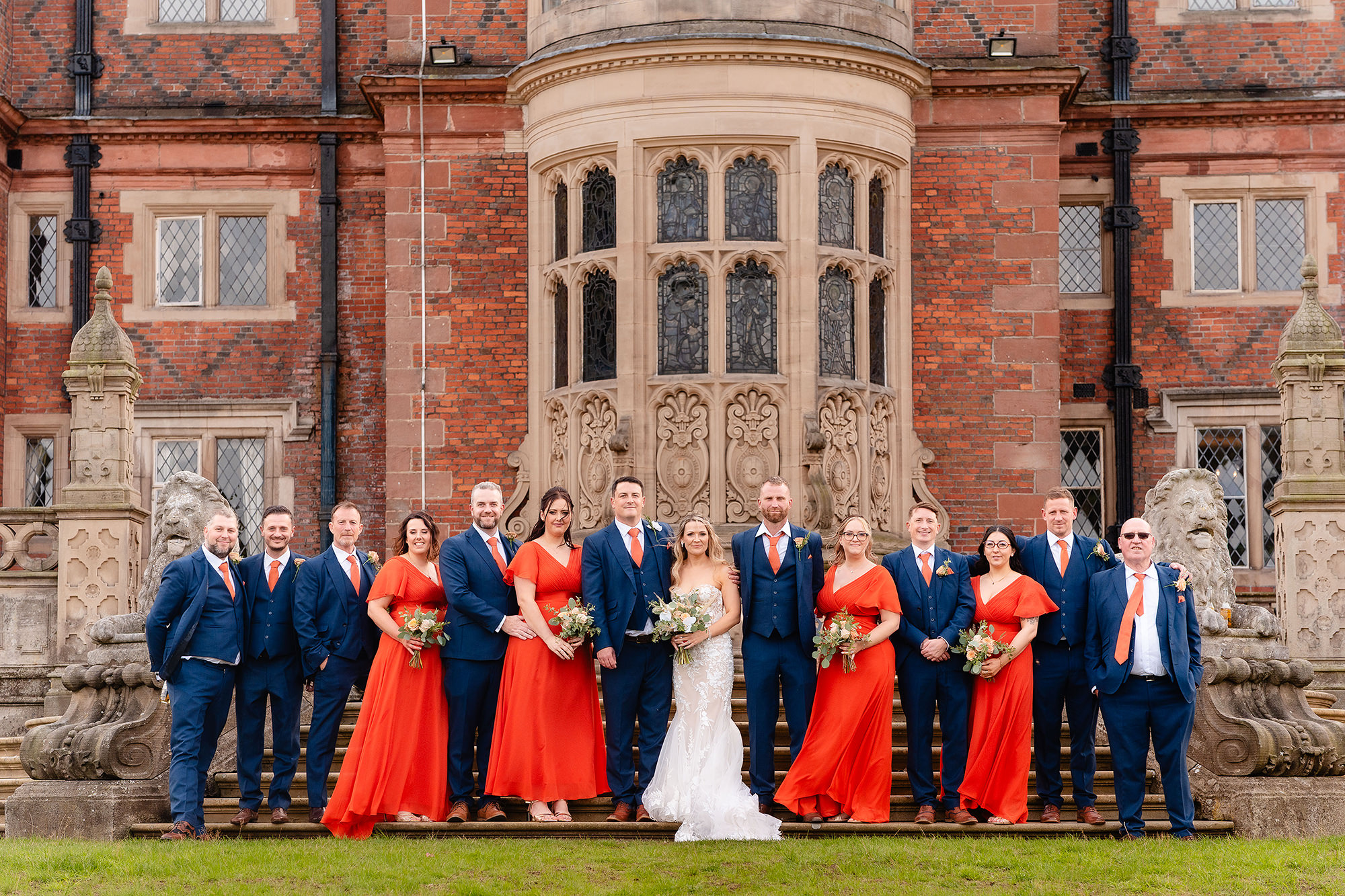 The full wedding party lined up on the steps at the back of Crewe Hall, navy suits and burnt orange bridesmaid dresses