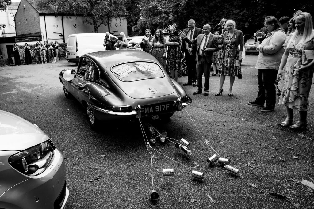 E-type Jaguar driving away from the church trailing tin cans as guests wave, black and white, Cheshire wedding photographer