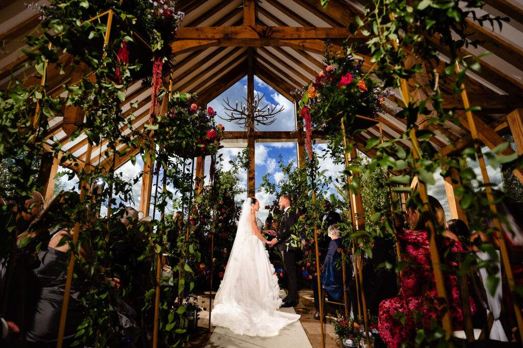 Wide view of the ceremony under hanging florals and the glass gable of the barn, Nottingham wedding photographer