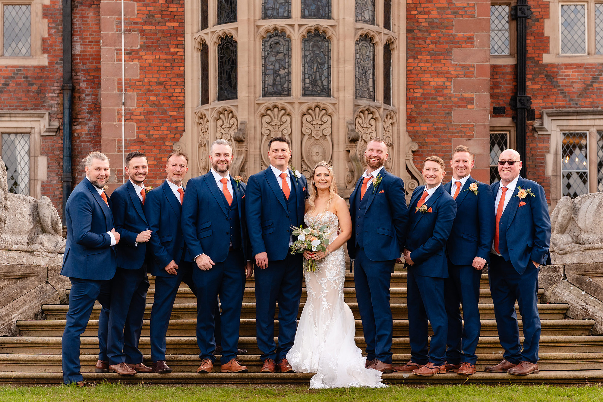 The bride and groom with a long line of men in navy suits and orange ties on the steps at the back of Crewe Hall