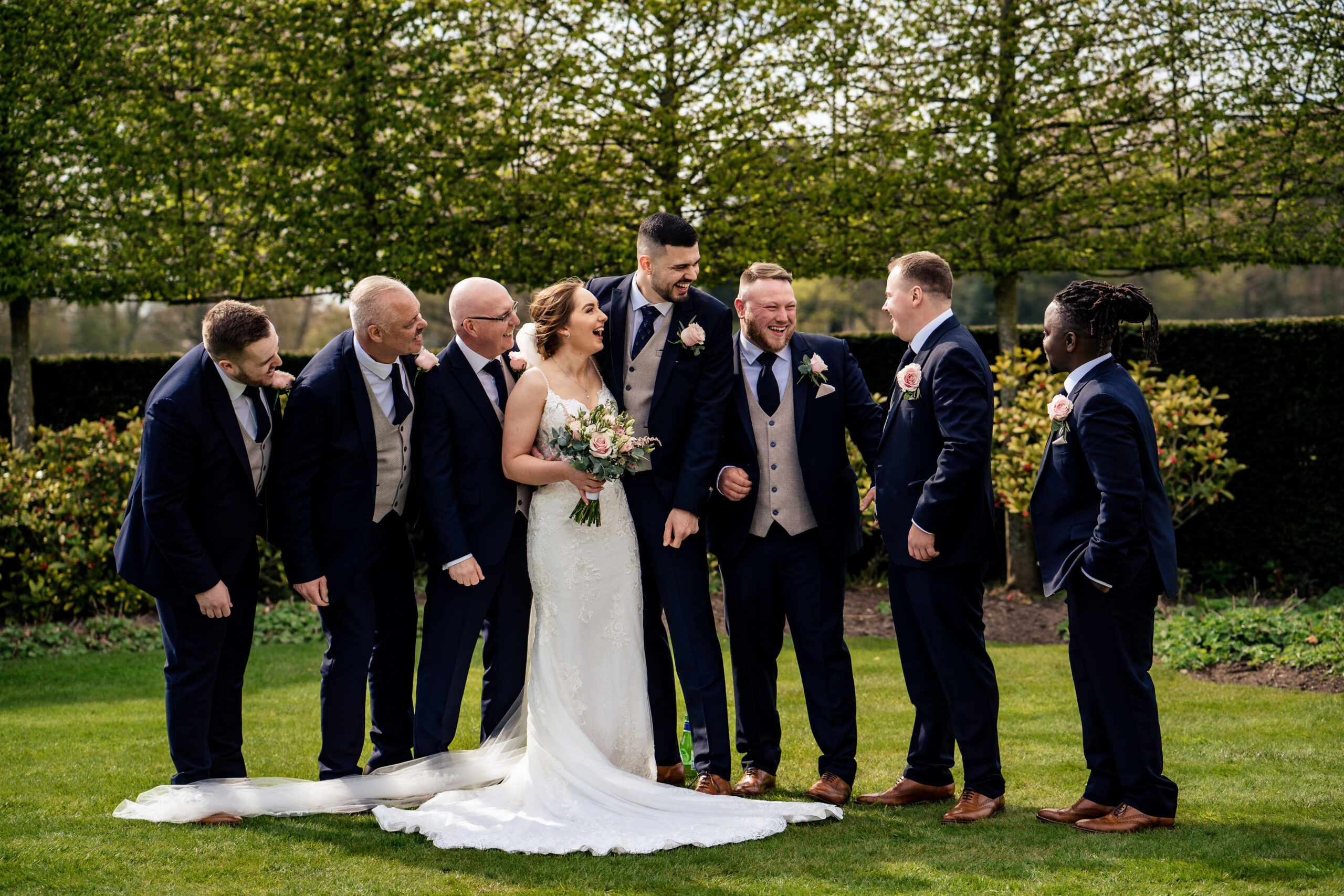 Bethany and Dean laughing with a group of men in navy suits on the lawn