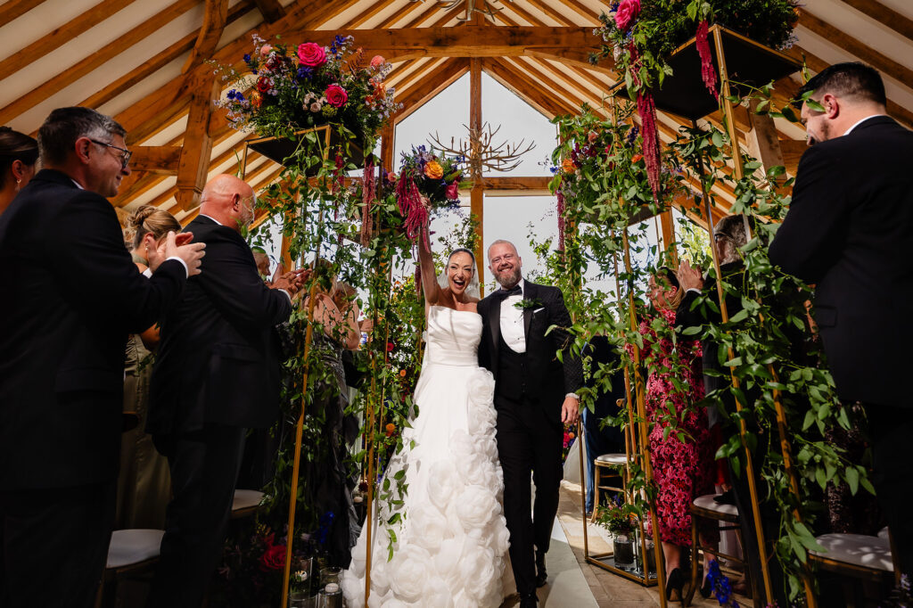 Kim throwing her arm in the air as she walks out with Glynn and guests applaud, Swancar Farm wedding photographer