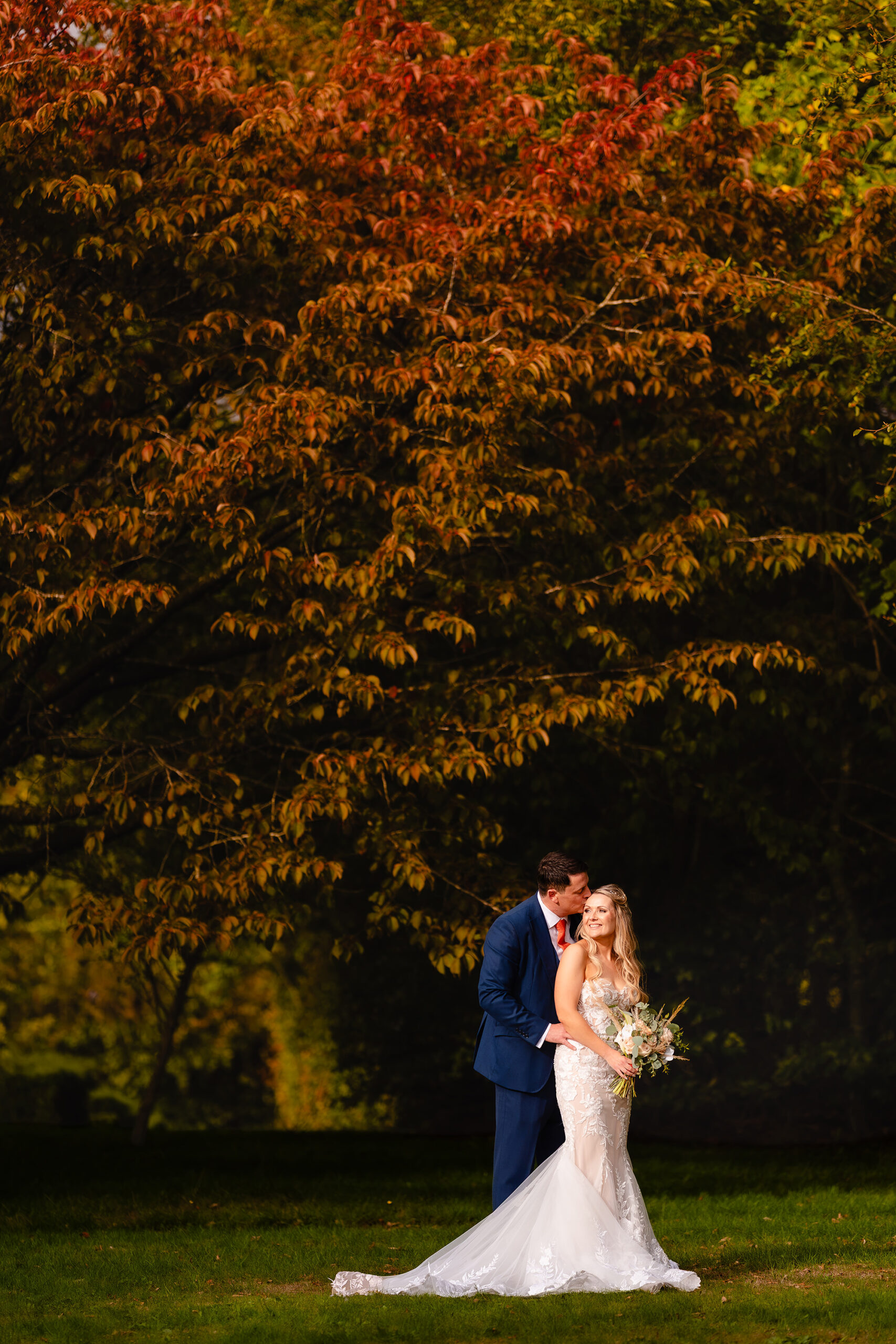 Bride and groom in an embrace on the lawn beneath a huge copper coloured tree in the grounds, Cheshire wedding photographer