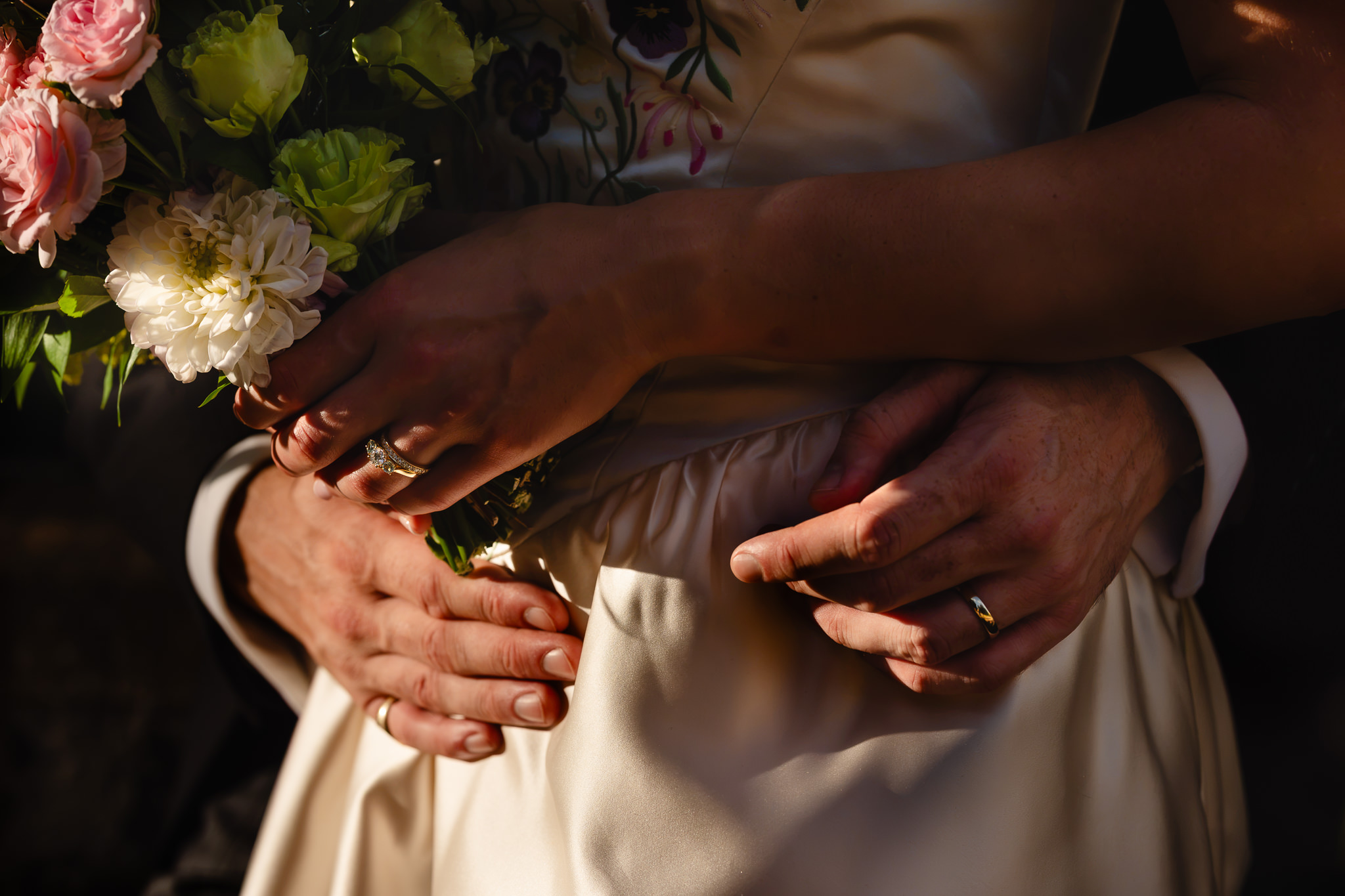 Close up of the couple’s hands and wedding rings resting against the bride’s silk dress
