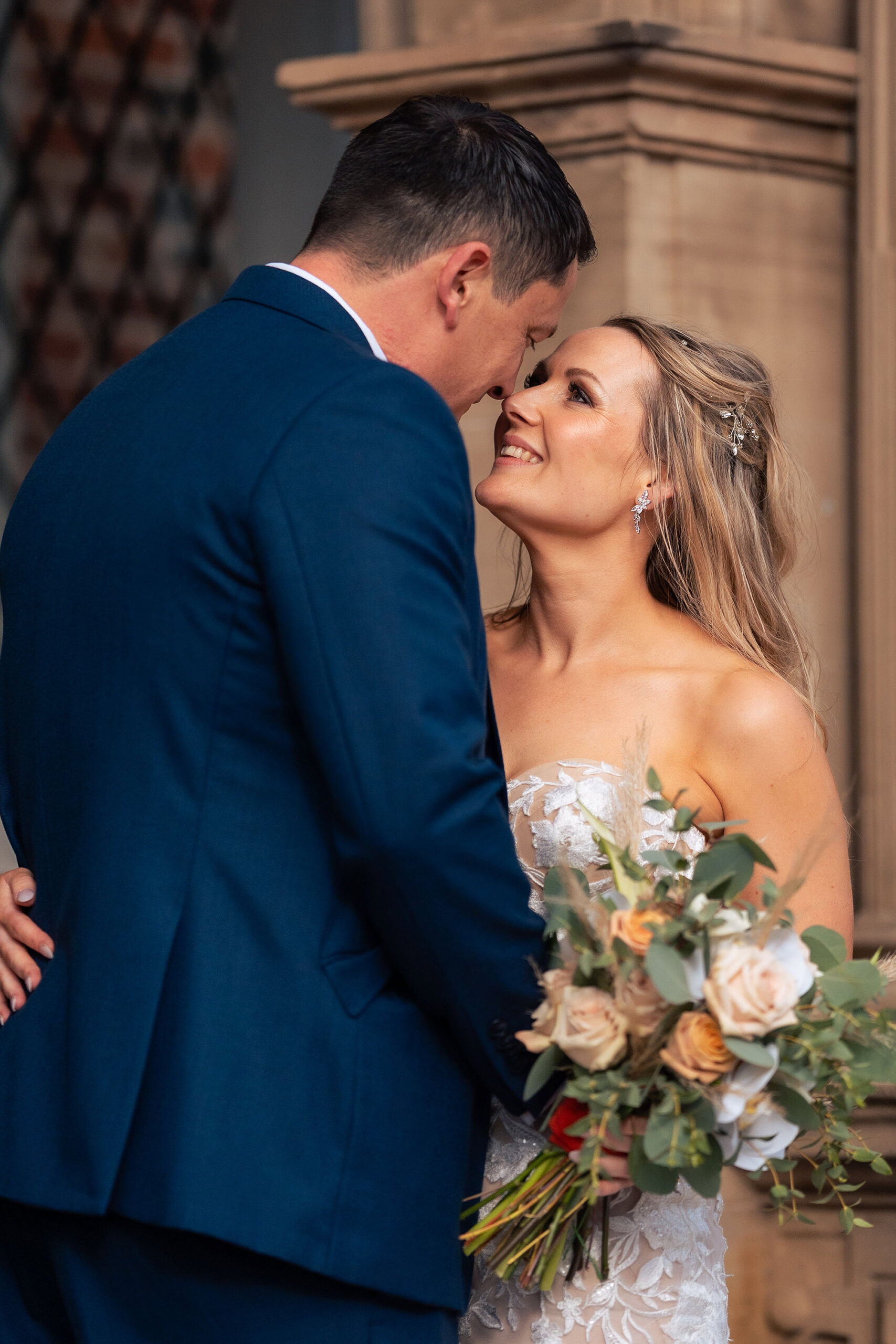 The couple nose to nose against the carved stonework of Crewe Hall, the bride holding her peach and eucalyptus bouquet