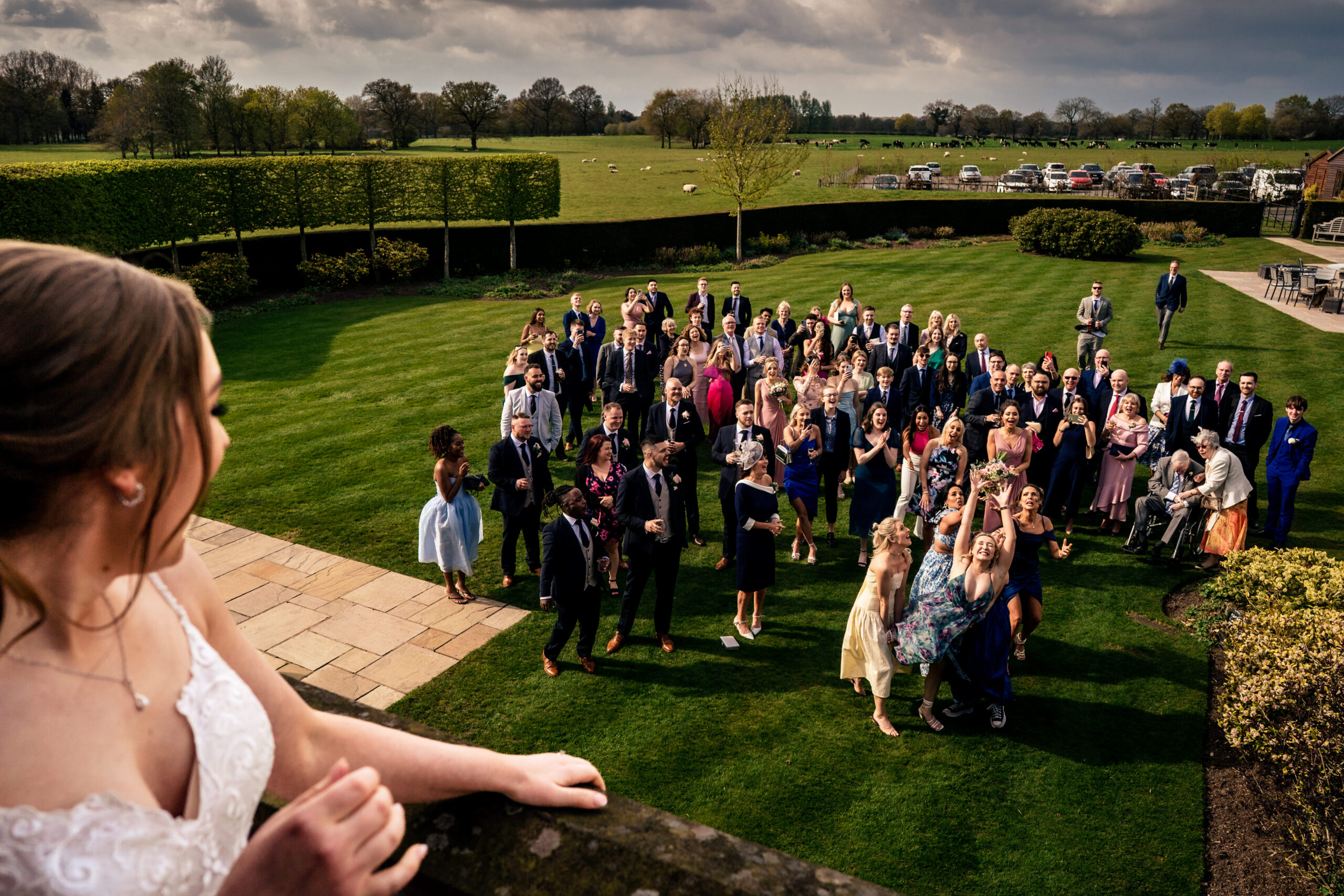 Bethany looking down from the balcony as guests on the lawn below reach for her bouquet
