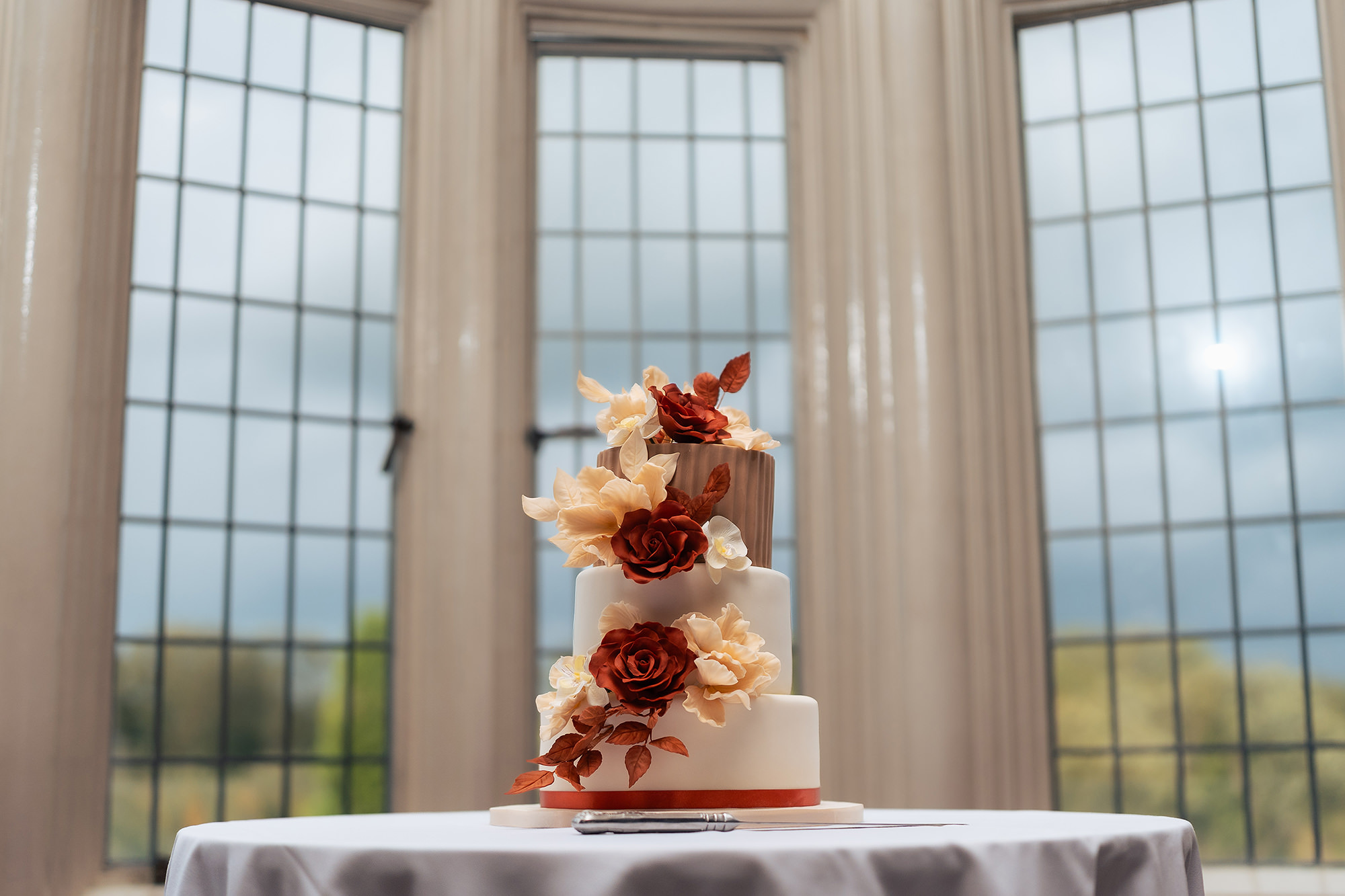 Three tier white wedding cake with rust and cream sugar roses and copper leaves, set in a bay window at Crewe Hall
