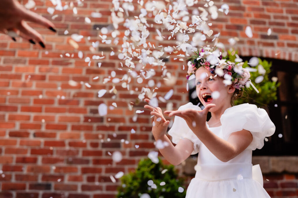 Flower girl throwing confetti with her mouth wide open against a red brick wall, Nottingham wedding photographer