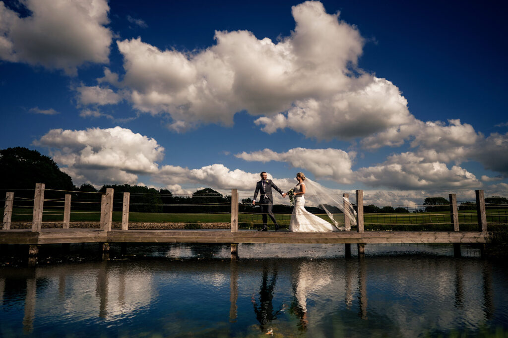 Bride and groom on the wooden jetty reflected in the lake at Merrydale Manor, veil lifting in the breeze, Cheshire wedding photographer