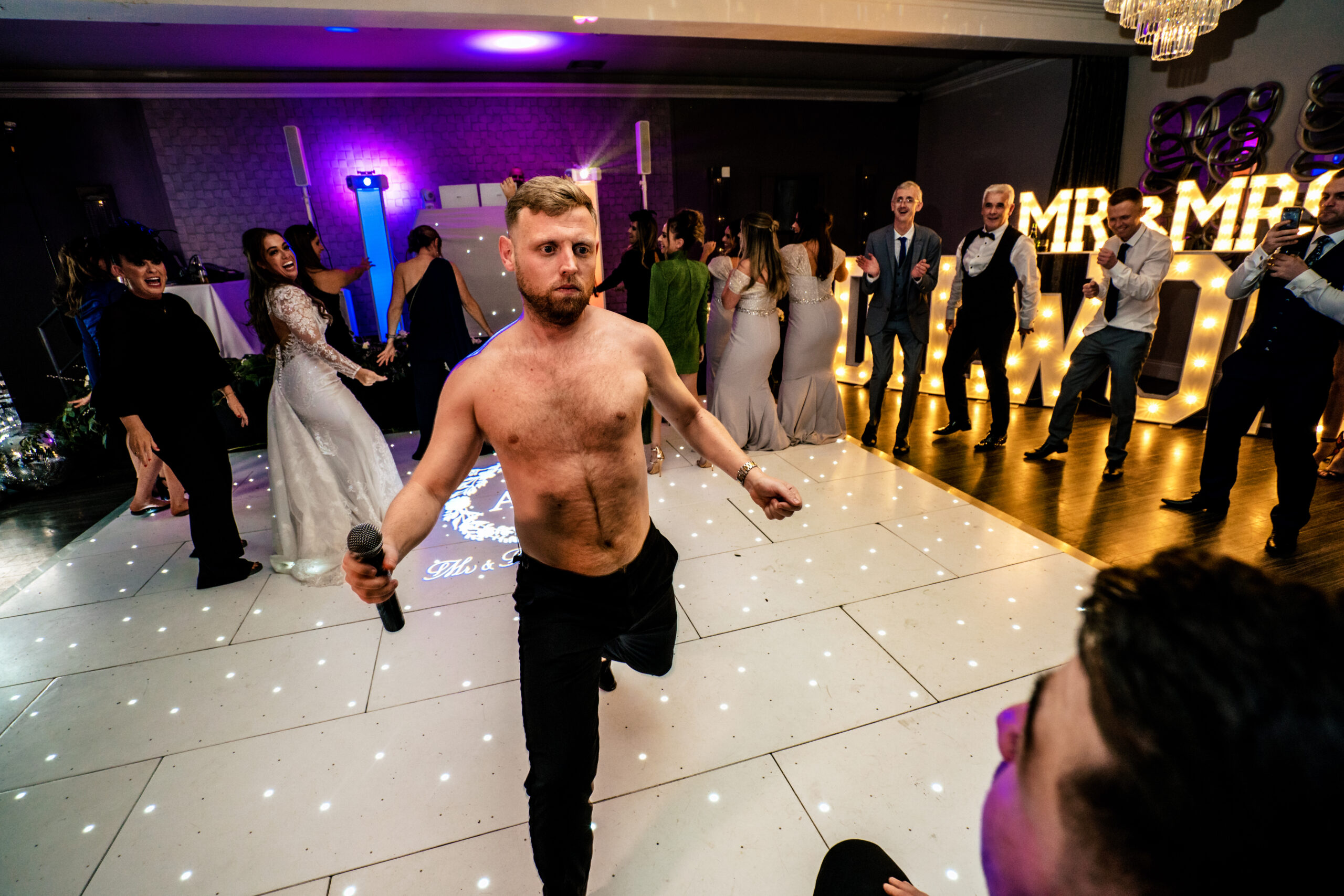 A shirtless man with a microphone mid dance on the dance floor at Merrydale Manor