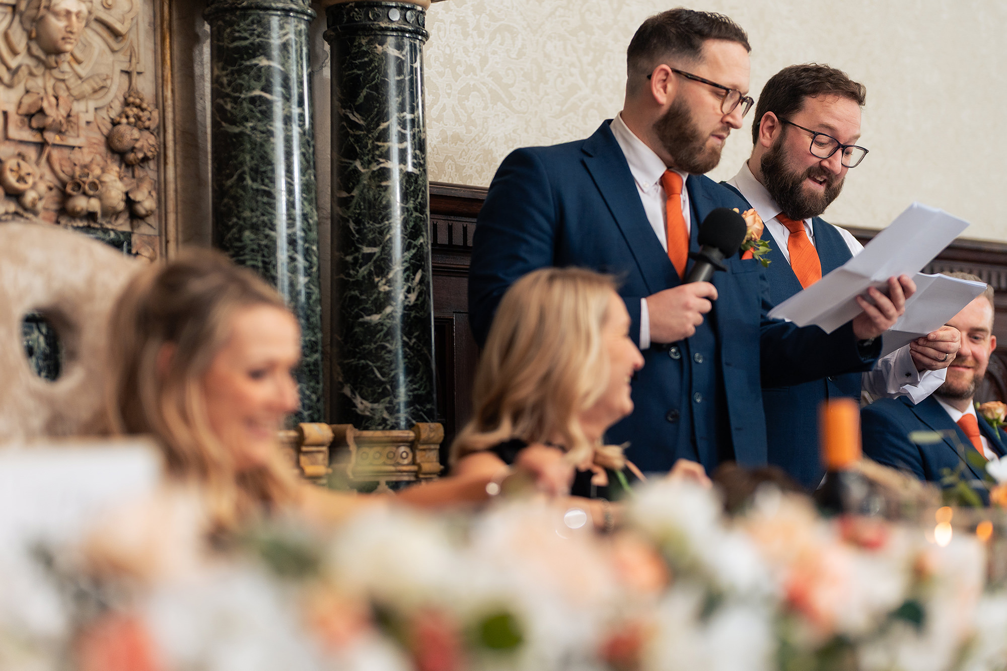 Two men in navy suits sharing a microphone and a sheet of paper as they deliver a speech in the Long Gallery