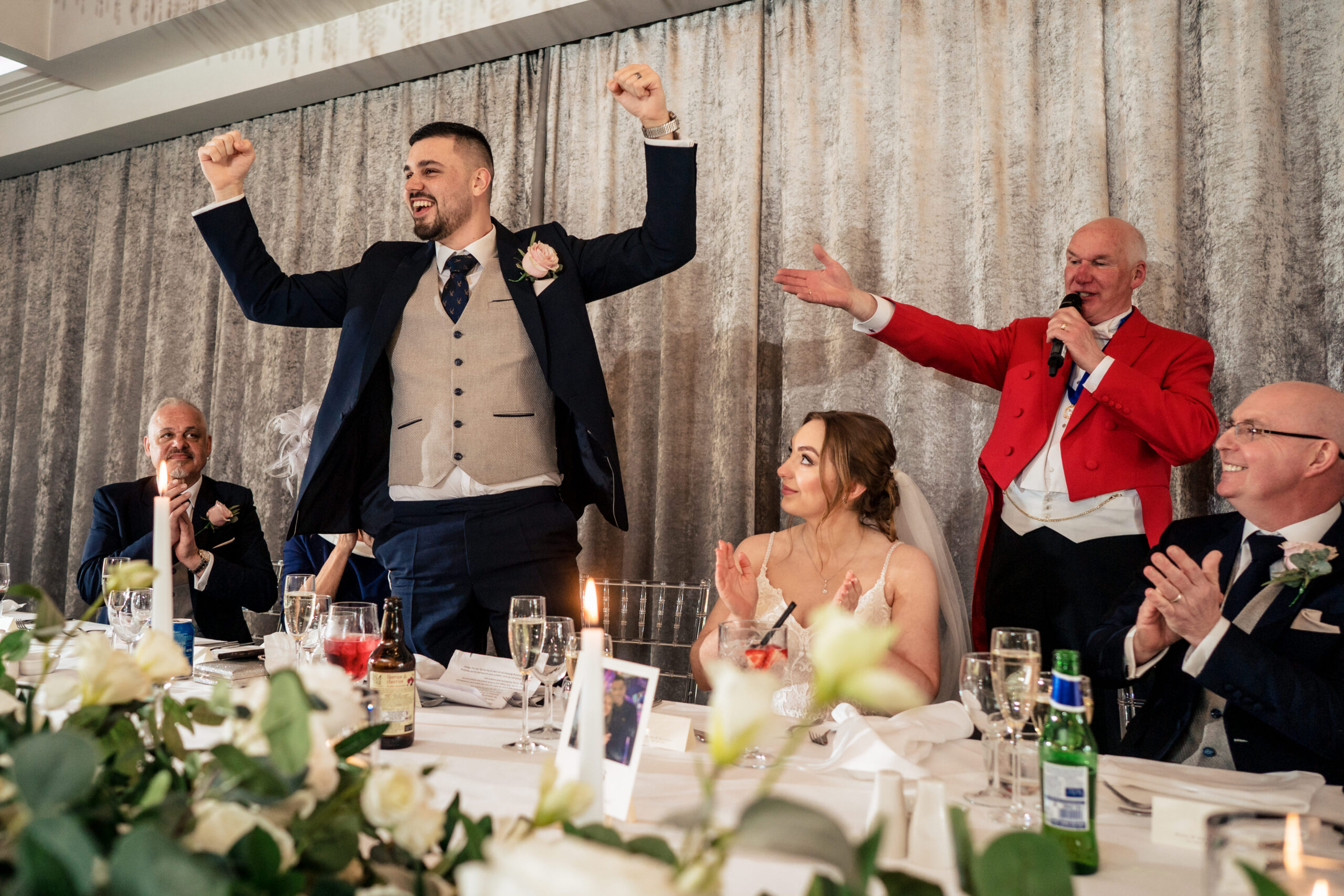 Dean standing with both arms in the air at the top table as Bethany laughs