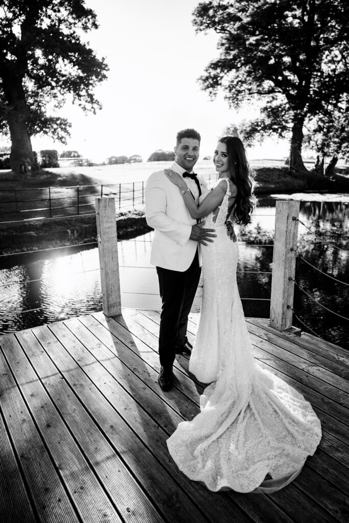 Black and white portrait of Gemma and Tyrone on the wooden jetty by the lake at Merrydale Manor, her train spread across the boards
