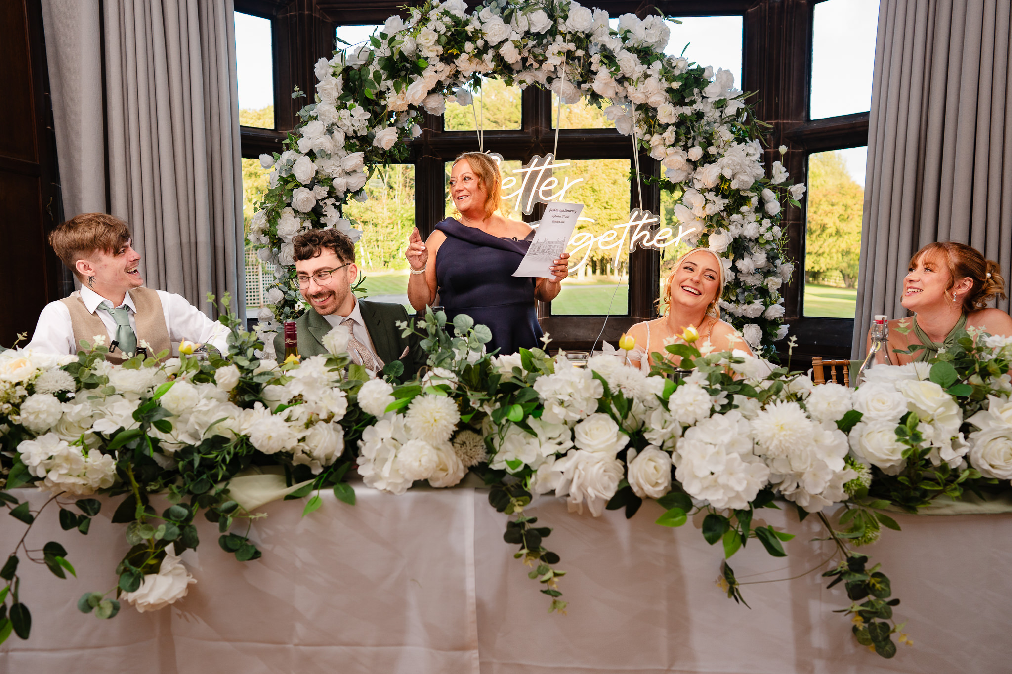 A speech being given at the top table, guests laughing along, framed by white floral displays and the Better Together sign