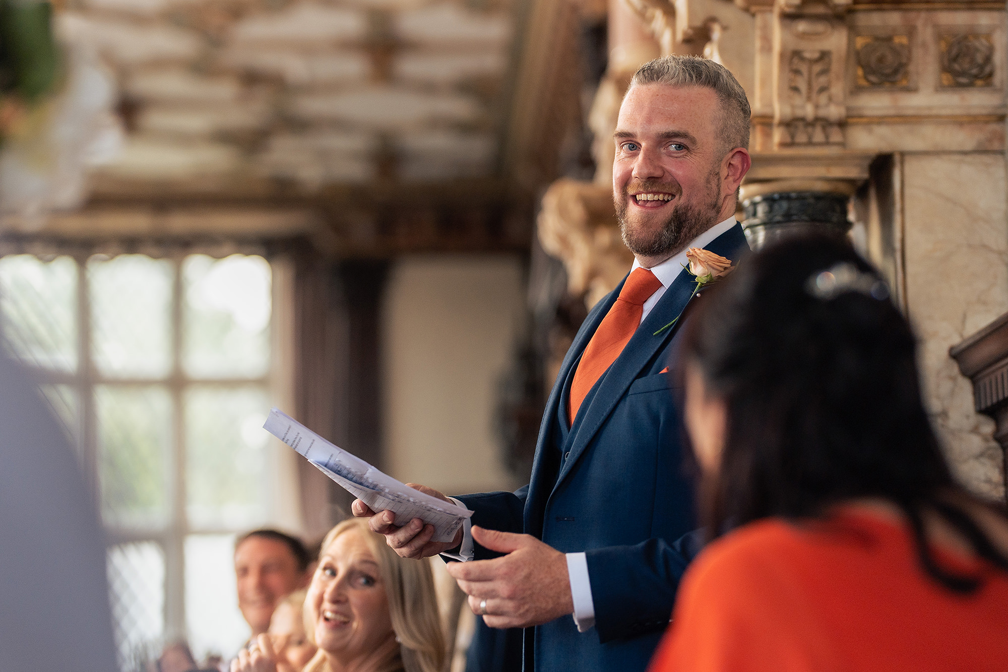 A man in a navy suit and orange tie grinning mid speech, notes in hand, in the Long Gallery at Crewe Hall