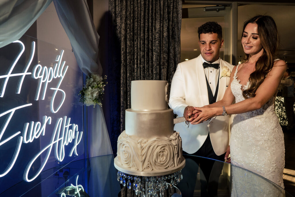 Gemma and Tyrone cutting their three tier white cake next to a Happily Ever After neon sign