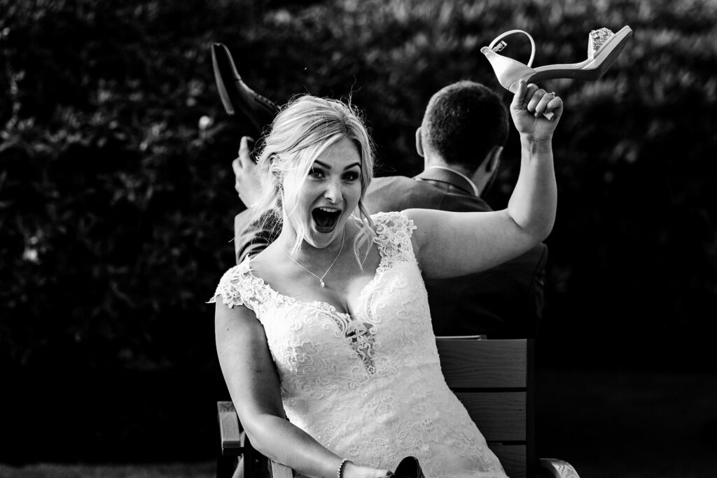 Bride laughing and holding up a pair of shoes during the shoe game, black and white, Cheshire wedding photographer