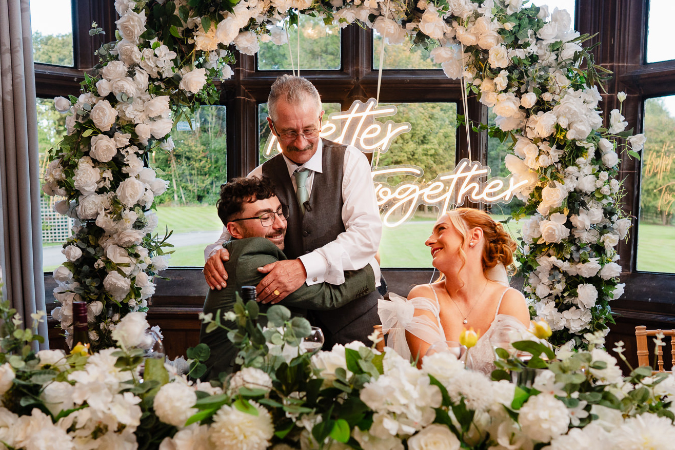 The bride’s dad hugging the groom at the top table, framed by the Better Together sign and white florals