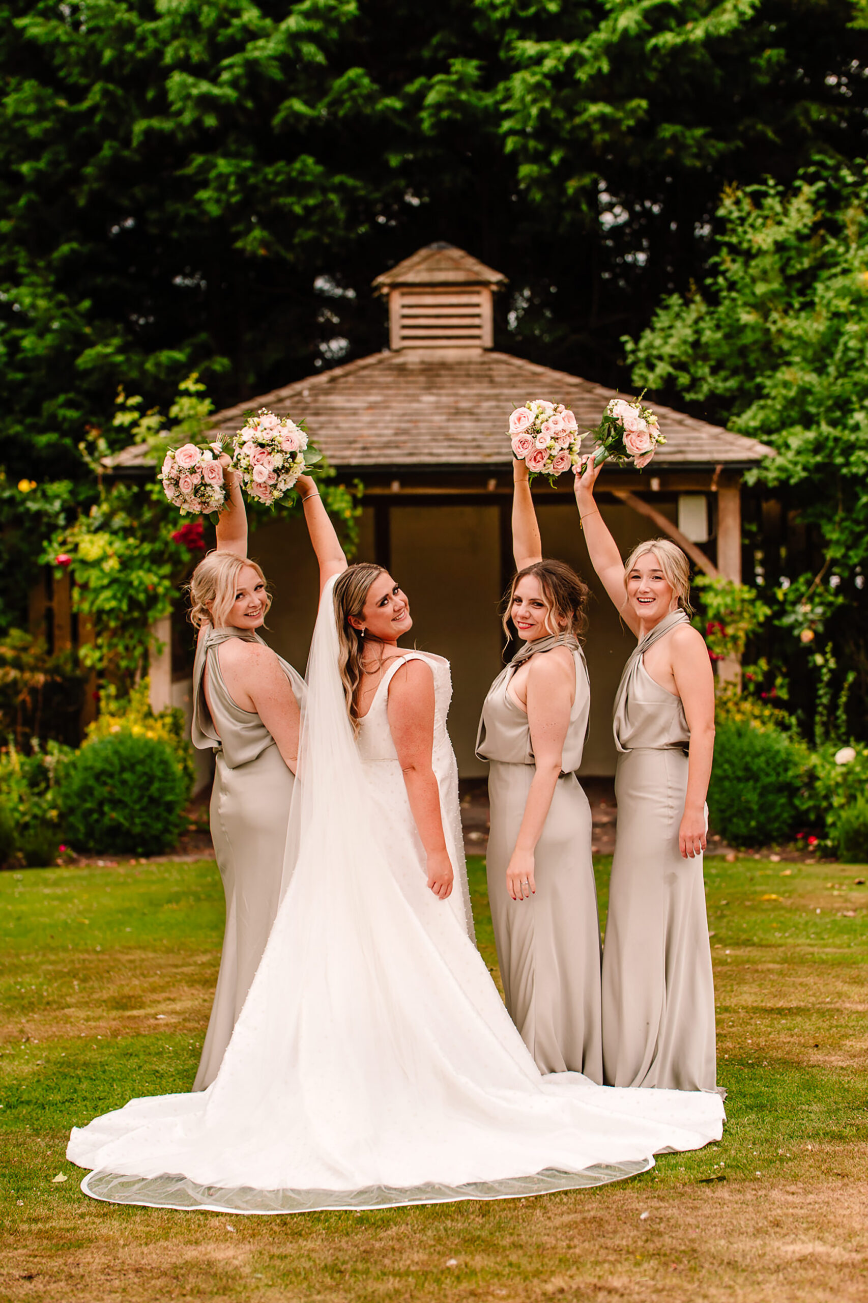 Chloe and her bridesmaids raising their bouquets and laughing by the gazebo at Wrenbury Hall
