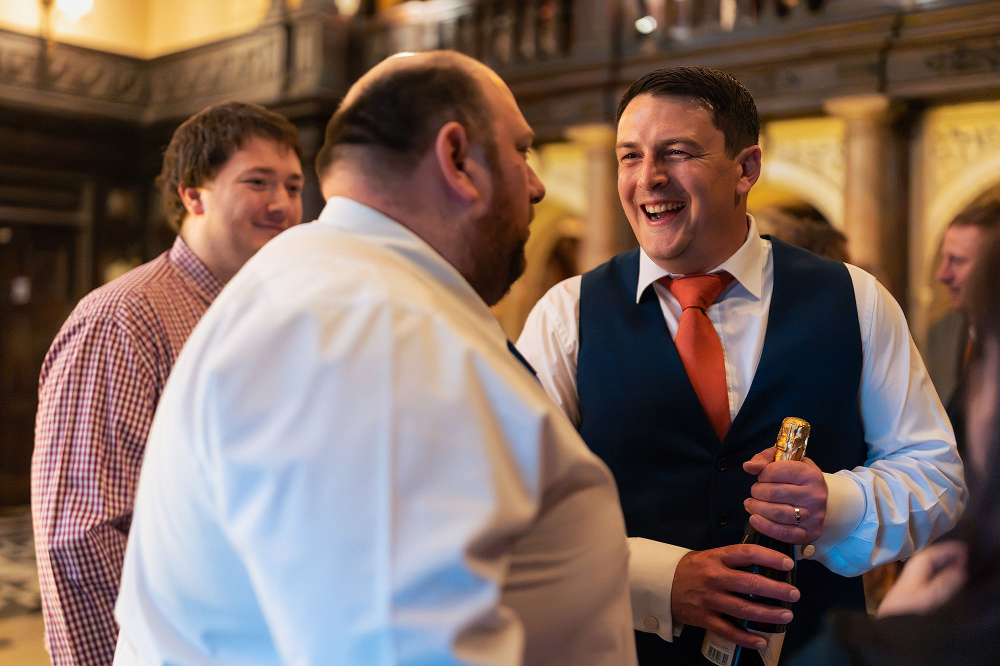The groom in his waistcoat and orange tie laughing with friends, bottle in hand, during the evening reception
